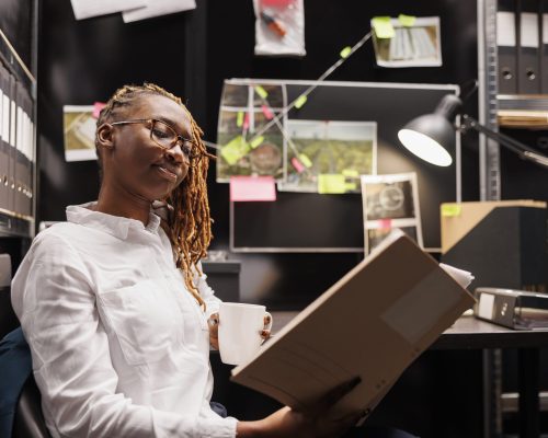 Policewoman reading investigation report while drinking coffee in office. African american woman police detective holding crime case file folder and analyzing evidence at night