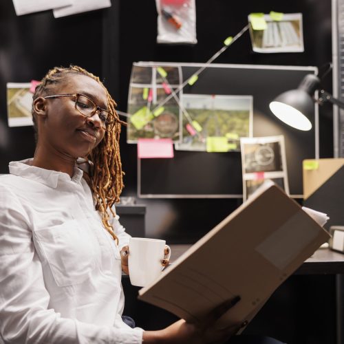 Policewoman reading investigation report while drinking coffee in office Policewoman reading investigation report while drinking coffee in office. African american woman police detective holding crime case file folder and analyzing evidence at night